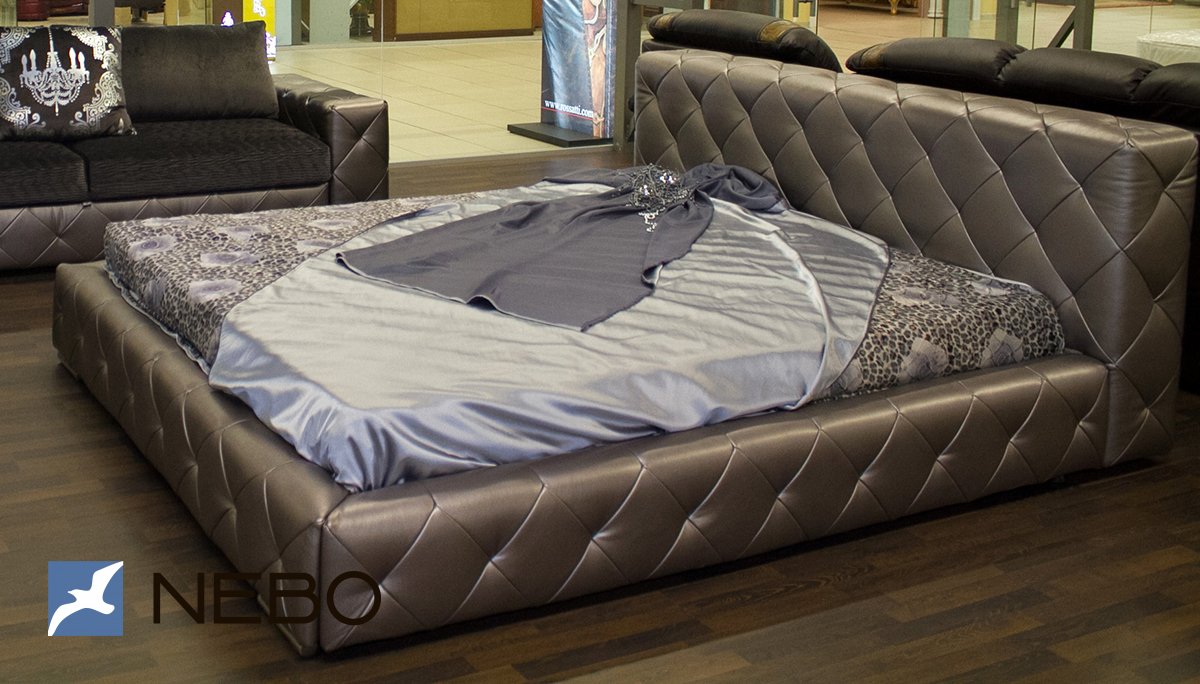 Мягкая кровать в обивке из серо-коричневой экокожи с пиковкой в классическом стиле