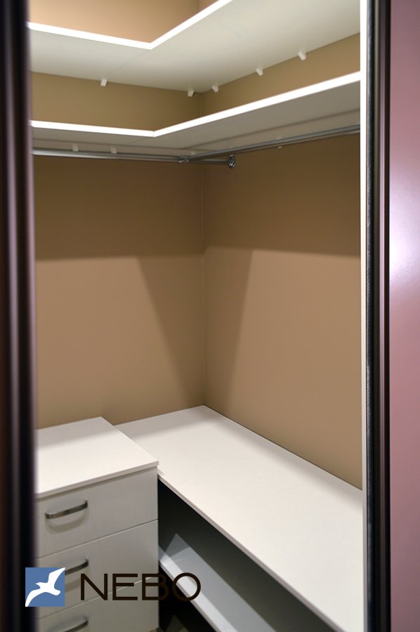 Маленькая гардеробная комната, меблированная белыми полками из ЛДСП, штангами для верхней одежды и тумбой