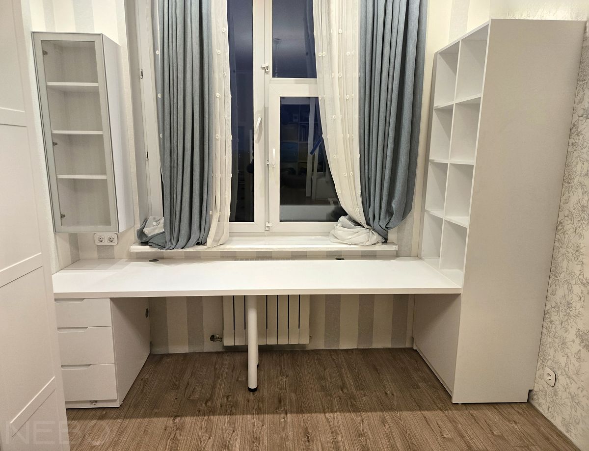 Белый стол у окна для двоих детей с навесным шкафчиком и стеллажом в детскую комнату